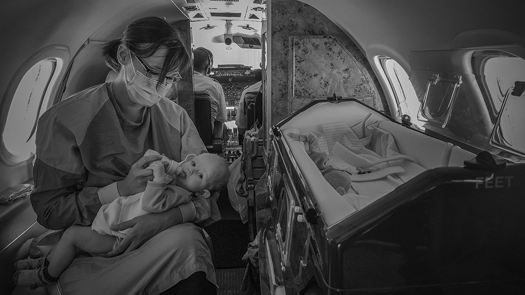 An intensive care nurse sits in an ambulance jet and gives a bottle of milk to a newborn patient.